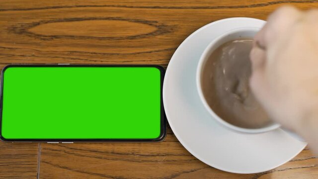 Phone with Horizontal Green Screen, Green Background, Chroma Key and Hand Stirring Hot Coffee, Closeup. Product Advertising, Business Concept, Video broadcast.