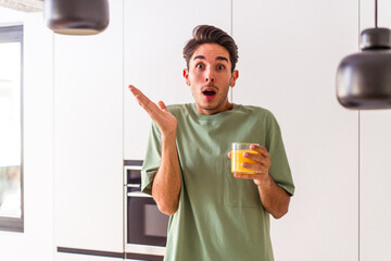 Young mixed race man drinking orange juice in his kitchen surprised and shocked.