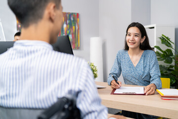 A job interview with a disabled man in wheelchair, beautiful woman of Asian Korean beauty asks questions to boy in shirt, she notes the answers, they are sitting in company office on two sides of desk