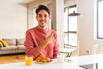 Young mixed race man having breakfast in a kitchen on the morning smiling and pointing aside, showing something at blank space.