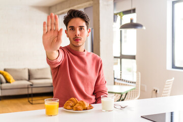 Young mixed race man having breakfast in a kitchen on the morning standing with outstretched hand showing stop sign, preventing you.