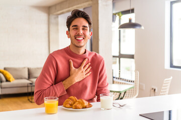 Young mixed race man having breakfast in a kitchen on the morning laughs out loudly keeping hand on chest.