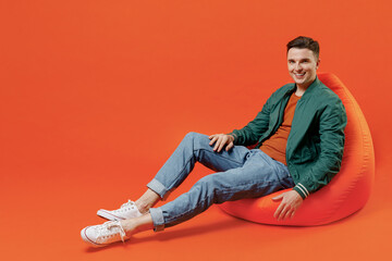 Full size body length happy fun young brunet man 20s wears red t-shirt green jacket sit in bag chair look camera isolated on plain orange background studio portrait. People emotions lifestyle concept.