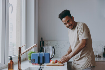 Young wondered african american man in casual clothes prepare sweet breakfast cut fruit watch series on mobile cell phone cooking food in indoor kitchen at home alone Healthy diet lifestyle concept