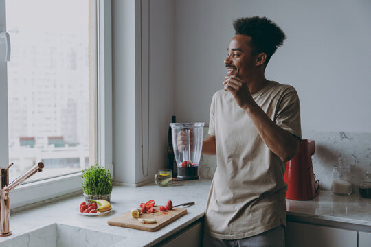 Young Smiling African American Man Wearing Casual Clothes Prepare Fruit Cocktail Eat Strawberry Using Mixer Blender Cooking Food In Light Indoor Kitchen At Home Alone. Healthy Diet Lifestyle Concept