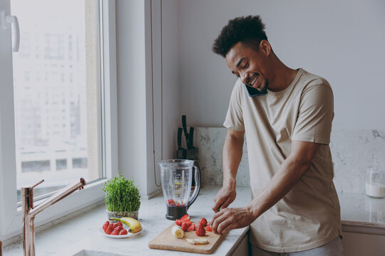 Young smiling happy african american man in casual clothes prepare fruit sweet breakfast talk speak on mobile cell phone cooking food in indoor kitchen at home alone Healthy diet lifestyle concept