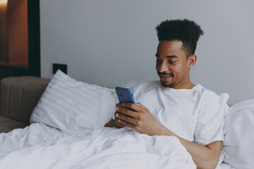 Young happy fun smiling african american man in nightwear use mobile cell phone lying in bed rest relax spend time in bedroom lounge home in own room house wake up dream be lost in reverie good day.