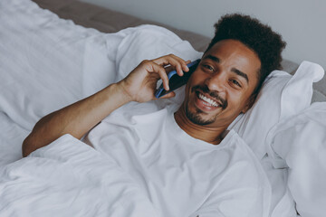 Young cheerful happy african american man in nightwear talk on mobile cell phone lying in bed rest relax spend time in bedroom lounge home in own room house wake up dream be lost in reverie good day