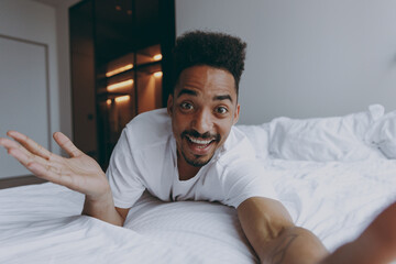 Fototapeta premium Close up young african american man in nightwear do selfie shot pov mobile phone spread hand lying in bed spend time in bedroom lounge home in own room house wake up dream be lost in reverie good day.