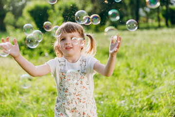 Little smiling cheerful happy kid girl 5-6 years old in white casual clothes blowing bubbles play on park green sunshine lawn, spending time outdoor in village countyside during summer time vacations.