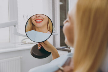 Young smiling satisfied happy woman 20s reflected in mirror sitting at dental office chair indoor light cabinet look at teeth toothy smile whitening result blink. Healthcare caries enamel treatment.