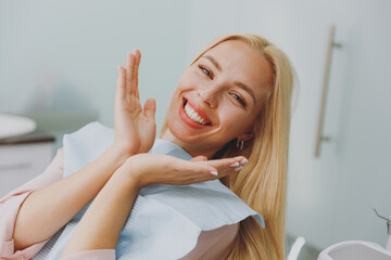 Close up young amazed smiling happy woman covered by napkin spread hands sit at dental office chair indoor light modern cabinet wait stomatologist for oral procedure Healthcare caries enamel treatment