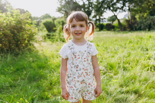 Little smiling happy beautiful kid girl 5-6 years old wear white casual clothes look camera play on park green sunshine lawn, spending time outdoor in village countyside during summer time vacations.