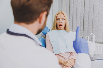 Young man dentist doctor in white gown talk speaking with patient consulting shocked scared european woman sit at dentist office chair indoor cabinet near stomatologist Healthcare enamel treatment