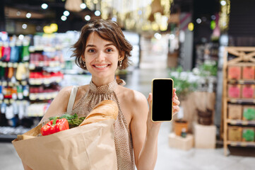 Young woman 20s in casual clothes shopping at supermaket store with craft paper package with groceries show mobile cell phone blank screen workspace area inside hypermarket. Gastronomy food concept