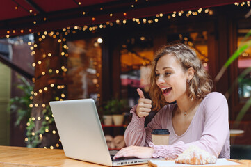 Young woman in casual clothes sit alone at table in cafe shop eat breakfast hold use work on laptop pc computer talk by viceo call show thumb up drink coffee relax in restaurant in free time indoors