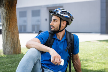 Side view of muslim deliveryman in protective helmet holding smartphone on lawn