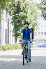 Fototapeta premium Happy arabian courier in safety helmet and backpack riding bike on urban street