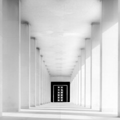 Photographie en noir et blanc d'une succession de colonnes en enfilade menant à une porte noire...