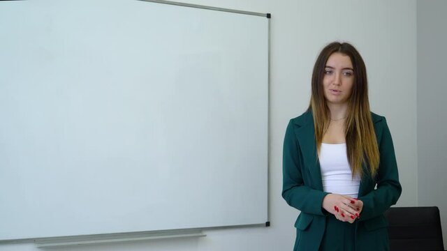 Young woman with long hair wearing green suit standing near whiteboard, gesturing and giving speech. Business coach motivating her audience. Concept of training