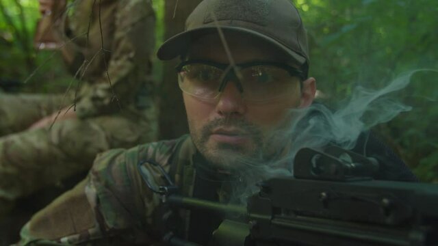 Close-up portrait of military watcher with machine gun letting smoke out of mouth, smoking while observing enemy territory in forest area, covering fellow soldiers during resting in halt outdoors