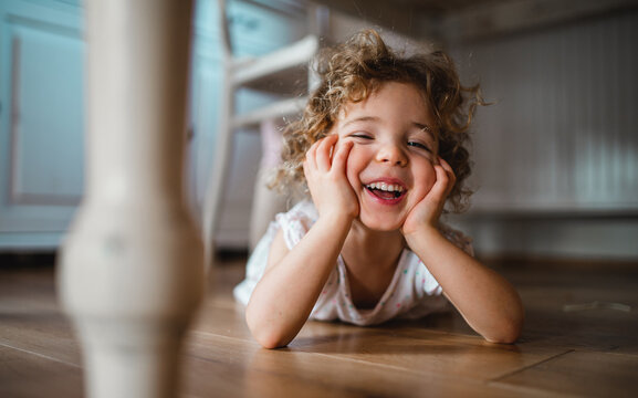 Portrait of cute small girl lying on floor under table indoors at home, looking at camera.