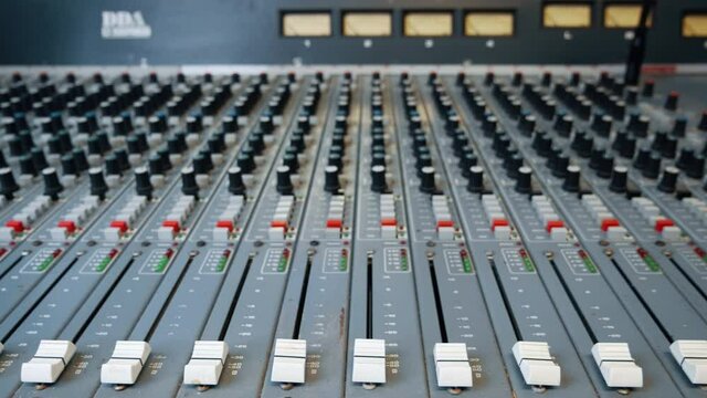 Vintage recording studio mixing desk faders. The music mixer console has faders in the foreground and pan pot knobs with VU meters in the background.