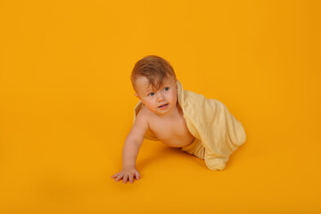 a beautiful little blue-eyed boy covered with a yellow towel crawls on a yellow background