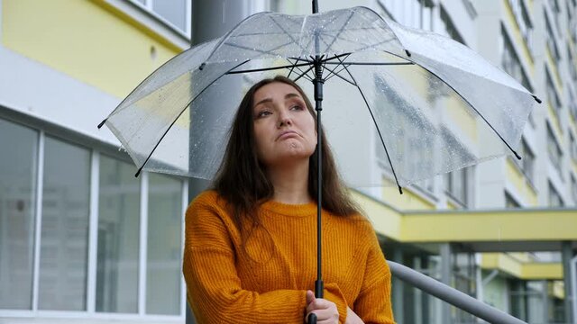 sad woman stands under a transparent umbrella in the rain on the background of an apartment building.