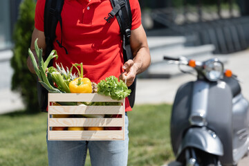 Cropped view of deliveryman in red t-shirt holding box with organic vegetables near blurred scooter - Powered by Adobe