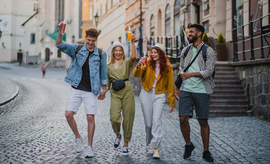 Front view of group of happy young people with drinks outdoors on street on town trip, laughing.