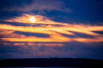 Evening river landscape with the setting sun. Calm water and dramatic sky with clouds. Trees grow on the nearest shore. There is a copy space