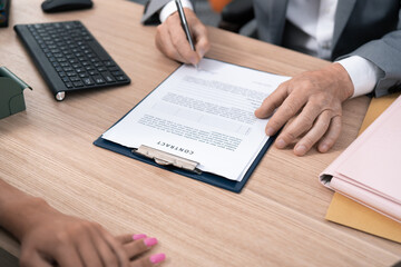 A document, an employment contract signed by the employer, boss, manager, hands of an elderly man signing on a piece of paper, sitting at a desk across from a female employee