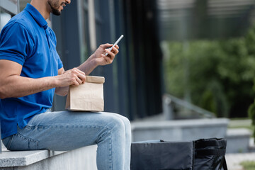 Cropped view of smiling courier using smartphone and holding paper bag outdoors