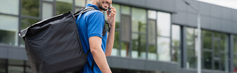 Cropped view of smiling deliveryman with thermo backpack talking on smartphone outdoors, banner