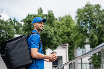Smiling arabian courier with thermo backpack holding coffee cups outdoors