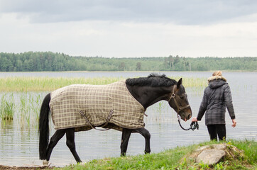 Young blonde woman walks on a horse in the lake water. The horse has a blanket on its back.