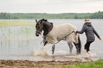 Young blonde woman walks on a horse in the lake water. The horse has a blanket on its back.