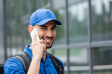 Positive arabian courier with backpack talking on smartphone outdoors