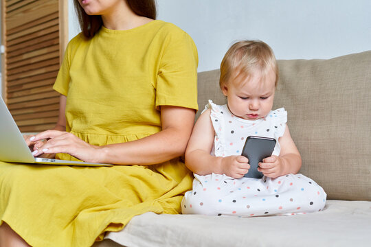 Faceless modern mother typing on computer while serious little toddler browsing cellphone. Concept of child mobile addiction and remote work challanges during quarantine