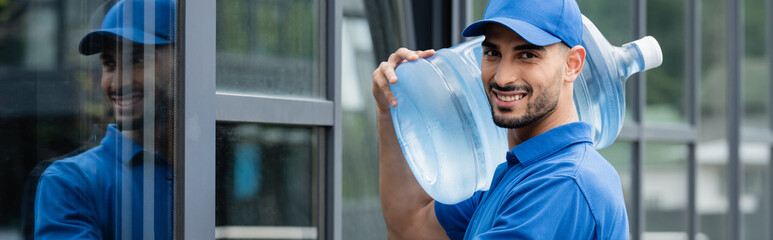 Smiling arabian courier holding bottle with water near building, banner