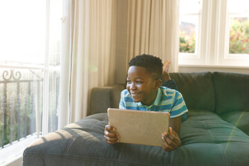 Smiling african american boy using tablet and lying on couch in living room - Powered by Adobe