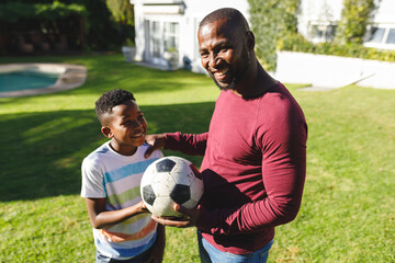 Portrait of smiling african american father with son embracing and holding football in sunny garden