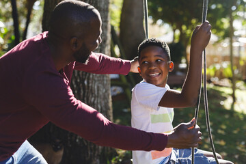 African american father with son having fun and playing on swing in garden