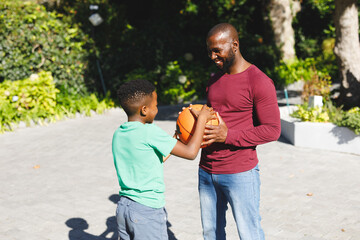 African american father with son smiling and playing basketball in garden