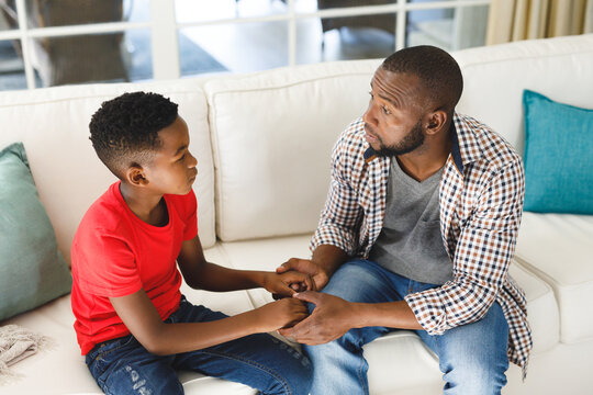 Serious African American Father And Son Sitting On Couch In Living Room Talking And Holding Hands