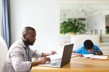 African american father working on laptop in dining room with son sitting with him doing homework