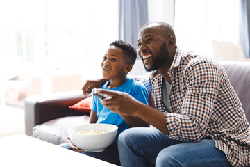 Happy african american father and son sitting on sofa, watching tv