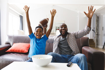 Excited african american father and son sitting on sofa, watching match on tv and cheering - Powered by Adobe