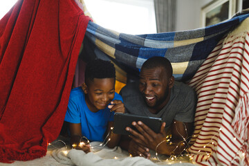 Happy african american father and son lying in blanket fort, using tablet - obrazy, fototapety, plakaty